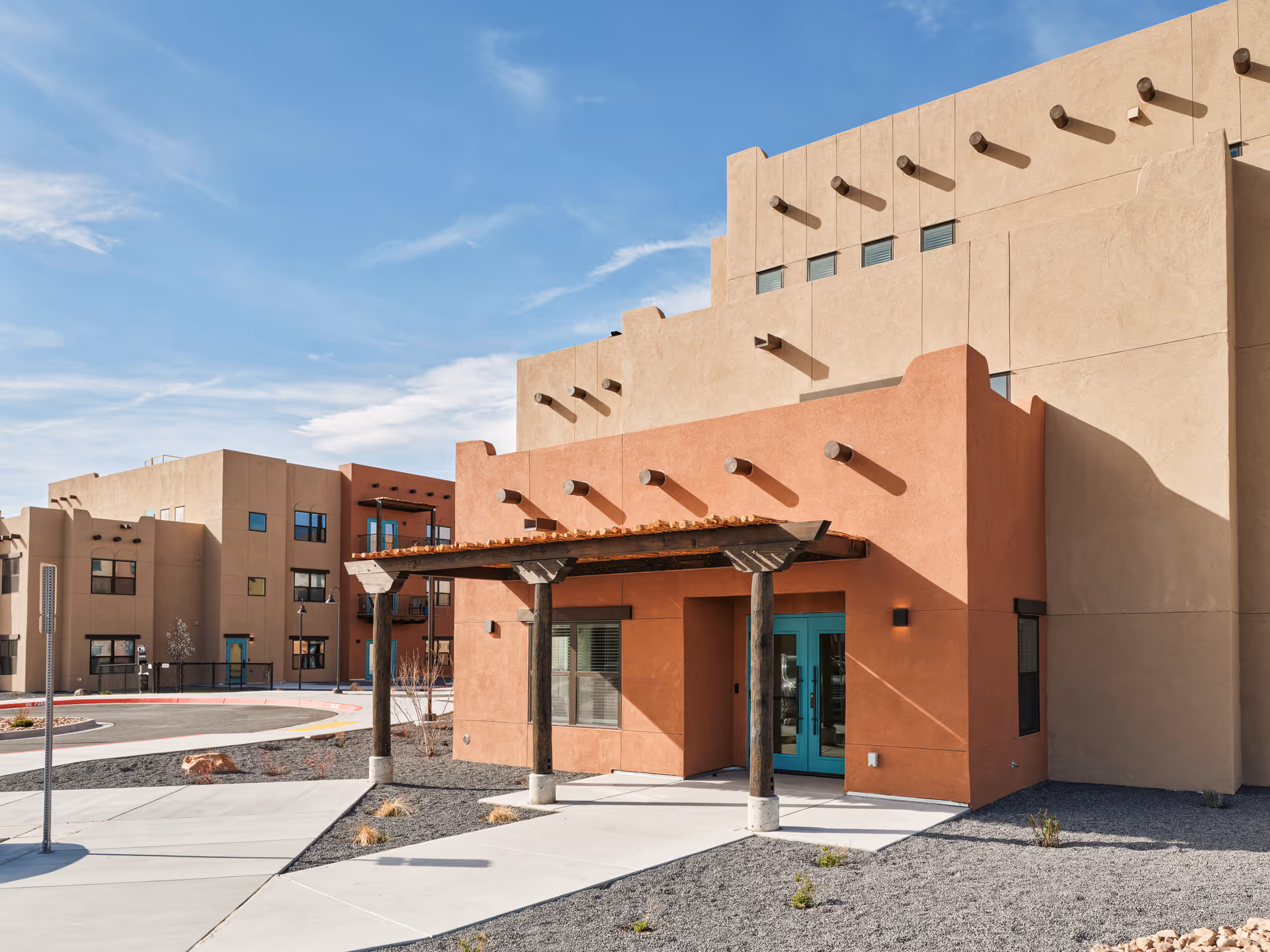 Exterior view of a senior living facility building with southwestern architectural style, featuring beige and terracotta stucco walls, wooden beams extending from the walls, a covered entrance with wooden posts, and a clear blue sky in the background.