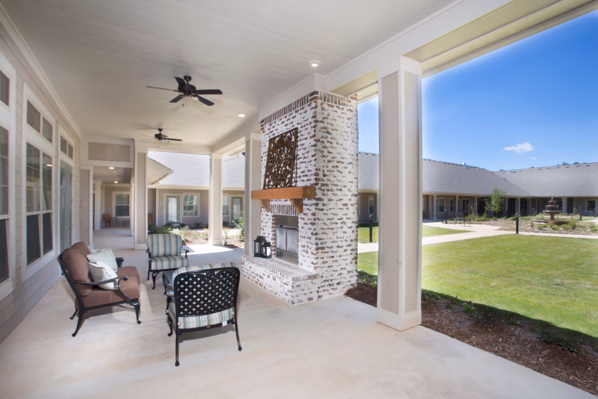 Covered outdoor patio with seating and a whitewashed brick fireplace overlooking a grassy courtyard and fountain.