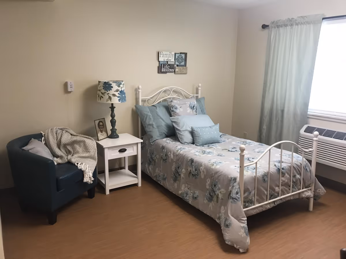 A cozy bedroom with a single white metal bed frame adorned with floral bedding and multiple pillows. Next to the bed is a white nightstand with a floral lamp and a framed black-and-white photo. A blue armchair with a gray pillow and a knitted throw blanket is positioned to the left of the nightstand. A window with light blue curtains and an air conditioning unit is on the right side of the room. A wall decoration above the bed reads 'Family is where Life Begins and Love Never Ends.'