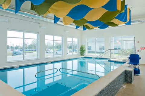 Indoor swimming pool area with large windows allowing natural light to fill the space. The pool has handrails and steps leading into the water. Colorful blue, green, and yellow fabric panels hang from the ceiling. There is a blue pool lift chair on the right side for accessibility.