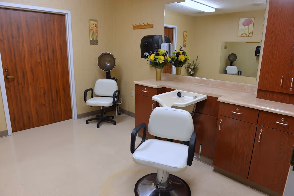 Interior of a small salon area with two white salon chairs, one positioned in front of a hair washing sink and the other under a vintage hair dryer. The room has wood-paneled cabinets, a large mirror, beige walls, a wooden door, and a vase of yellow and purple flowers on the counter.