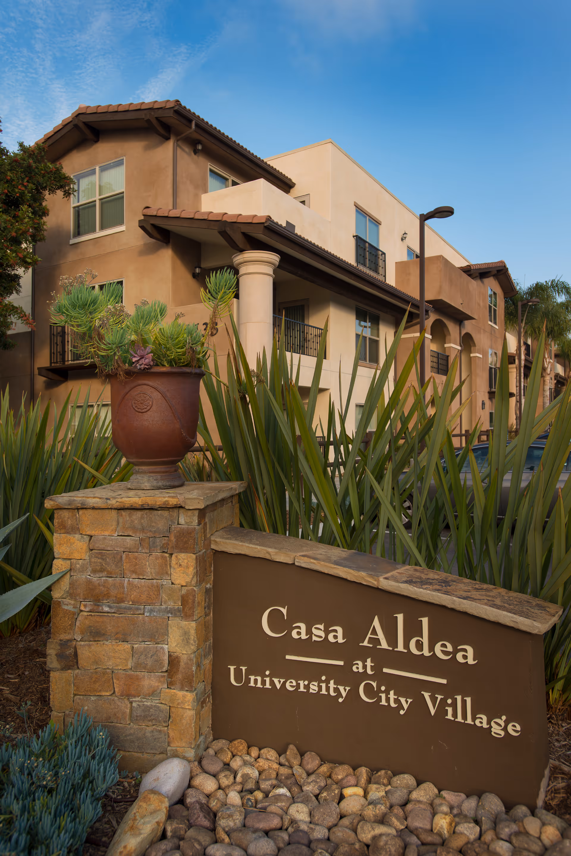 Exterior view of Casa Aldea at University City Village, showing a multi-story residential building with beige and brown walls, balconies, and a tiled roof. In the foreground, there is a stone sign with the facility's name and a large planter with green succulents. The sky is clear and blue.