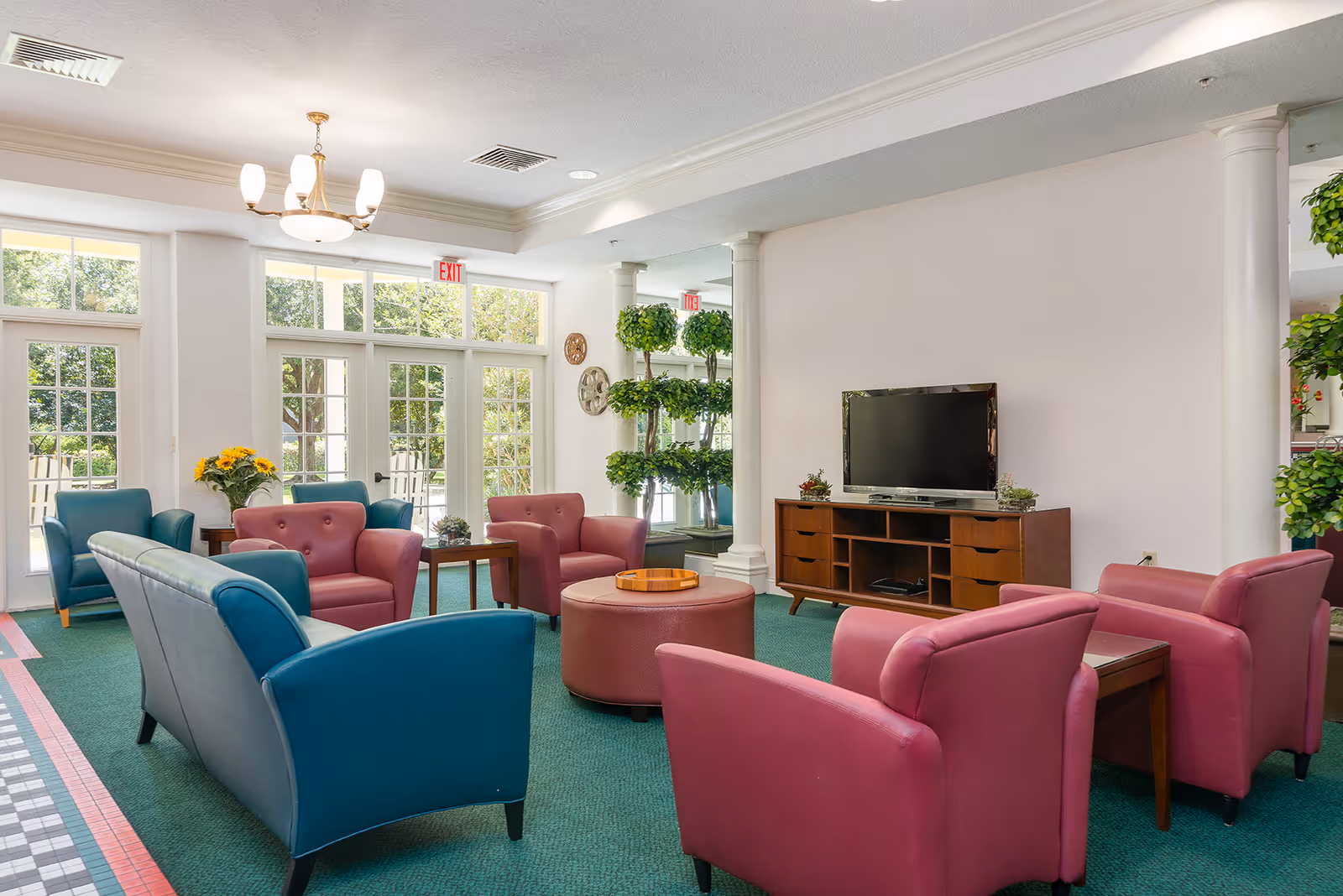 Bright lounge with colorful armchairs and sofas arranged around a central ottoman and a TV console, with large windows showing greenery outside.