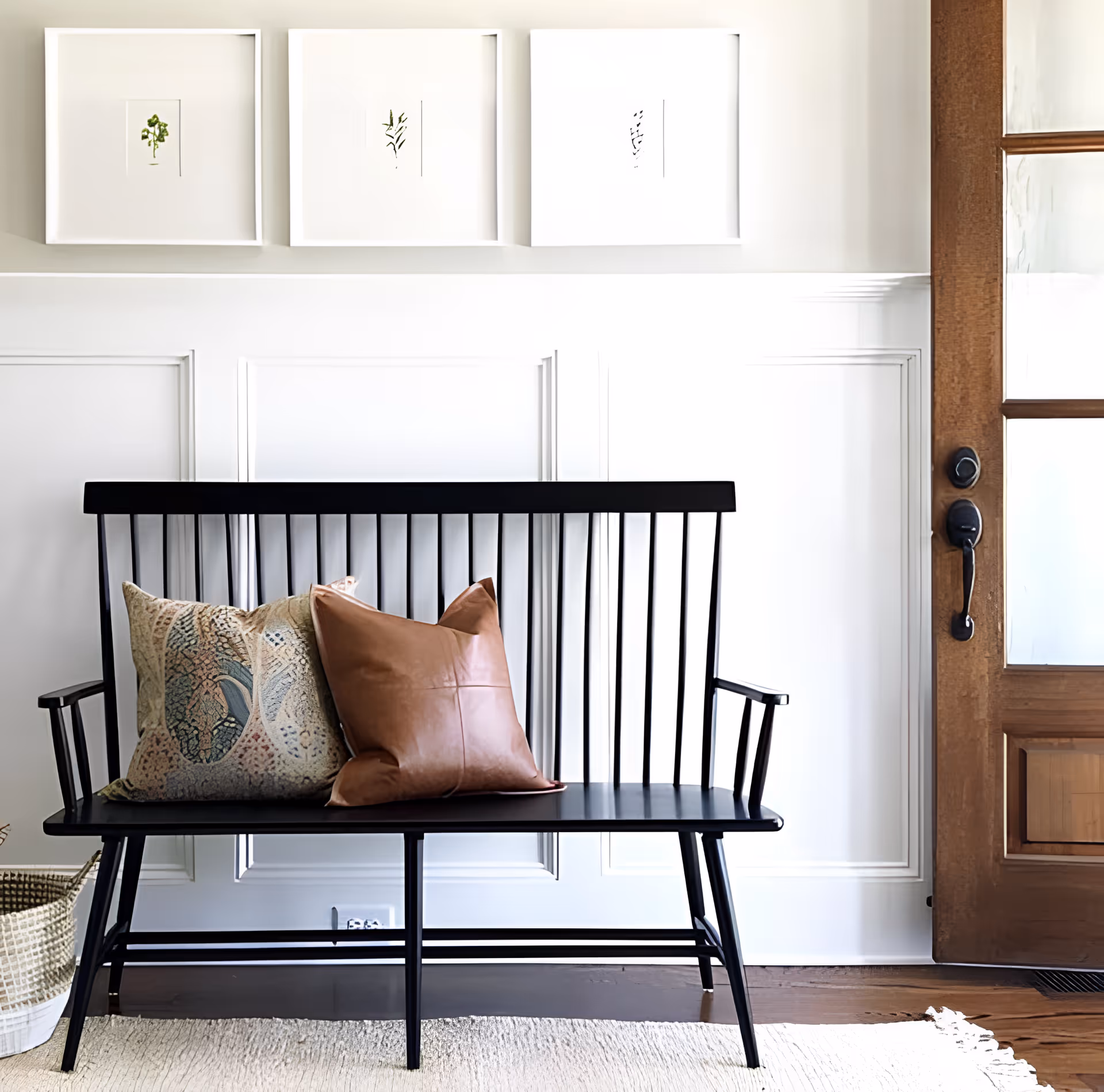 A black wooden bench with two decorative pillows, one patterned and one brown leather, placed against a white paneled wall. Above the bench are three framed botanical prints. To the right is a wooden door with glass panels, and a woven basket is partially visible on the left side on a light-colored rug.
