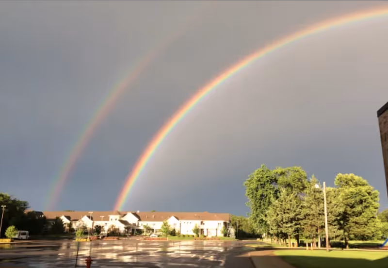 A double rainbow arches over a senior living facility building with a wet parking lot in the foreground and green trees on the right side under a cloudy sky.