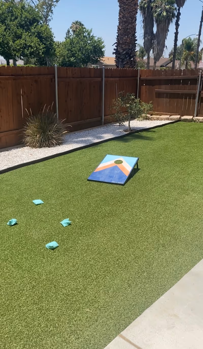 Outdoor area with artificial grass featuring a cornhole game board and four bean bags scattered on the ground. The area is enclosed by a wooden fence with some plants and palm trees visible in the background under a clear sky.
