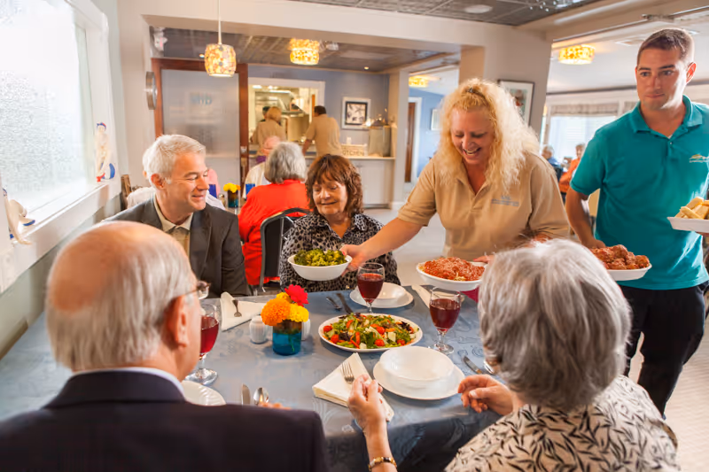 A group of elderly people sitting around a dining table with plates, glasses of red juice, and a salad bowl. Two staff members are serving bowls of food including salad and meatballs. The setting is a bright dining room with other people visible in the background.