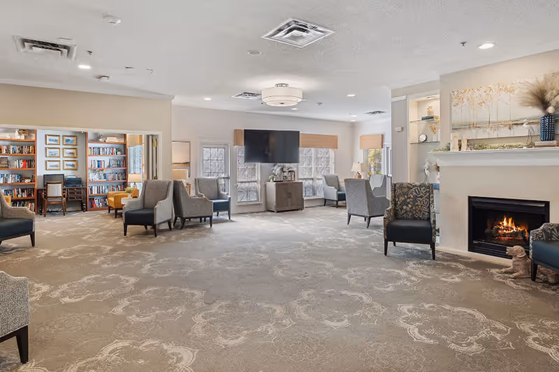 A spacious and well-lit living room area in a senior living facility with multiple comfortable armchairs arranged around the room. There is a fireplace with a small dog statue in front of it on the right side, a flat-screen TV mounted on the wall in the center, and a bookshelf-filled room visible through an open doorway on the left. The room features large windows with beige blinds, neutral-colored walls, and a patterned carpet.