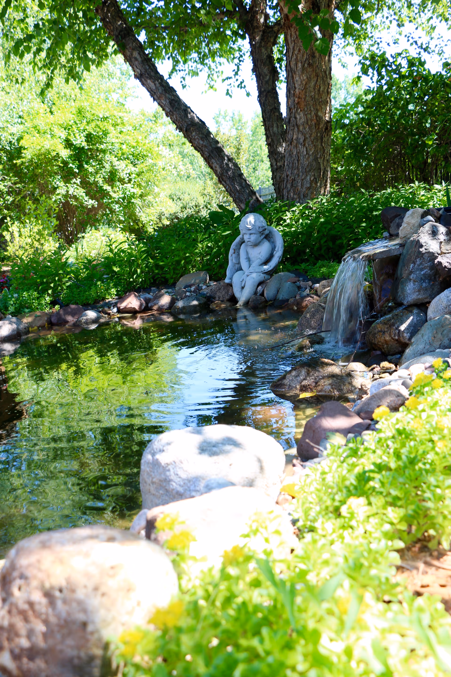 A serene garden pond surrounded by rocks and lush green plants with a small waterfall flowing into the pond. A statue of a seated angel is placed near the water's edge under a large tree.