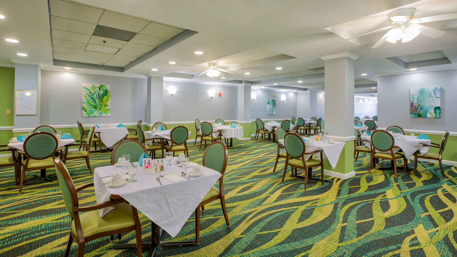 A spacious dining room with multiple tables covered in white tablecloths, each set with cups, glasses, napkins, and silverware. The chairs have green upholstery and wooden frames. The room features green and yellow patterned carpet, light gray walls with green wainscoting, ceiling fans, recessed lighting, and colorful abstract paintings on the walls.