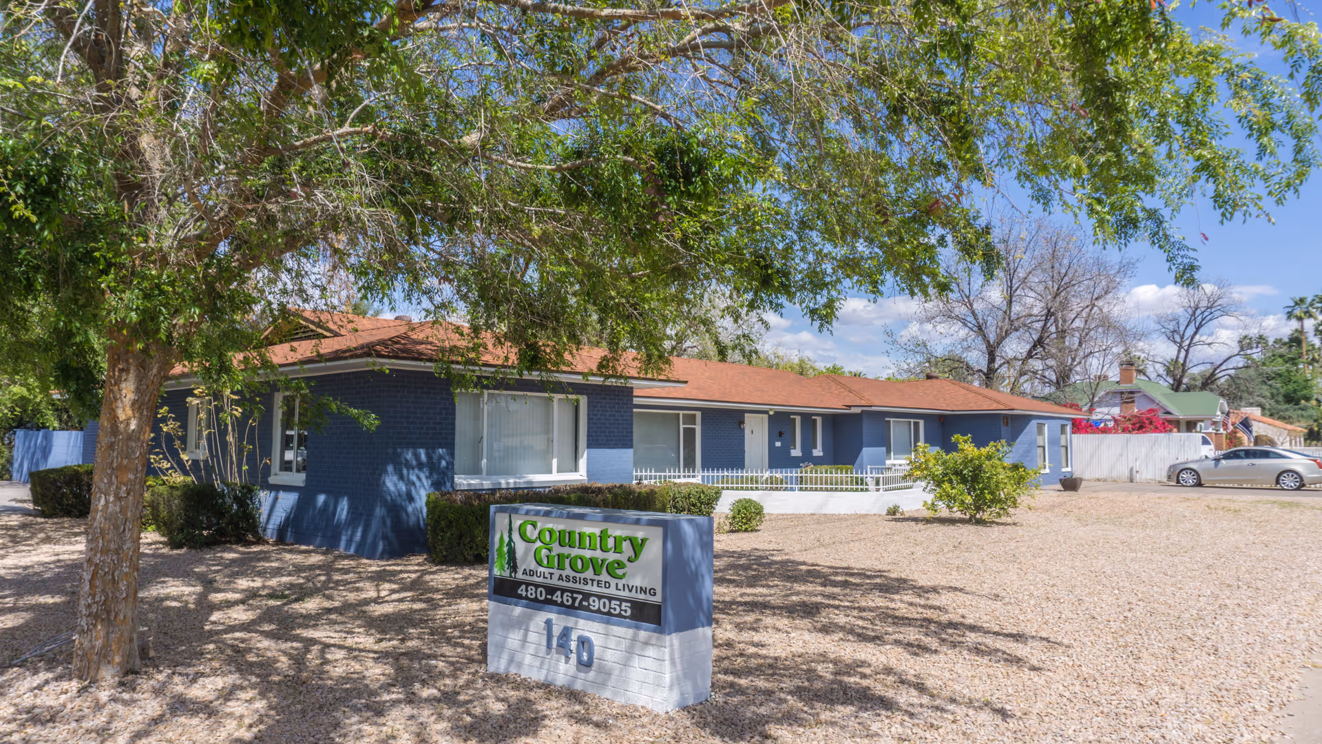 Exterior view of Country Grove Assisted Living Home, a single-story blue building with a red roof surrounded by trees and a gravel yard. A sign in front displays the facility name and phone number.