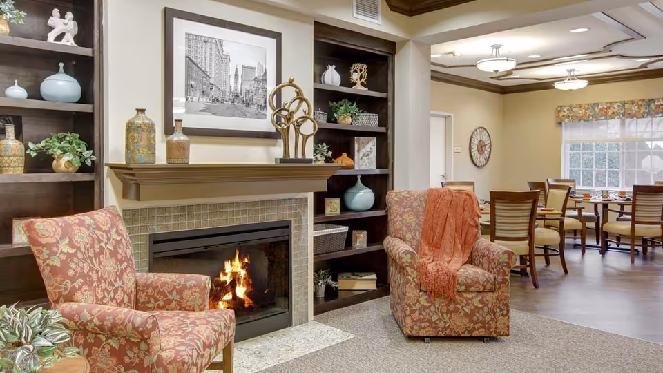 A cozy living area in a senior living facility featuring two floral upholstered armchairs, one with an orange throw blanket, positioned near a lit fireplace with a mantel decorated with vases and a sculpture. Behind the chairs are built-in dark wood shelves with decorative items and plants. In the background, there is a dining area with wooden chairs and tables set near a window with floral valances.