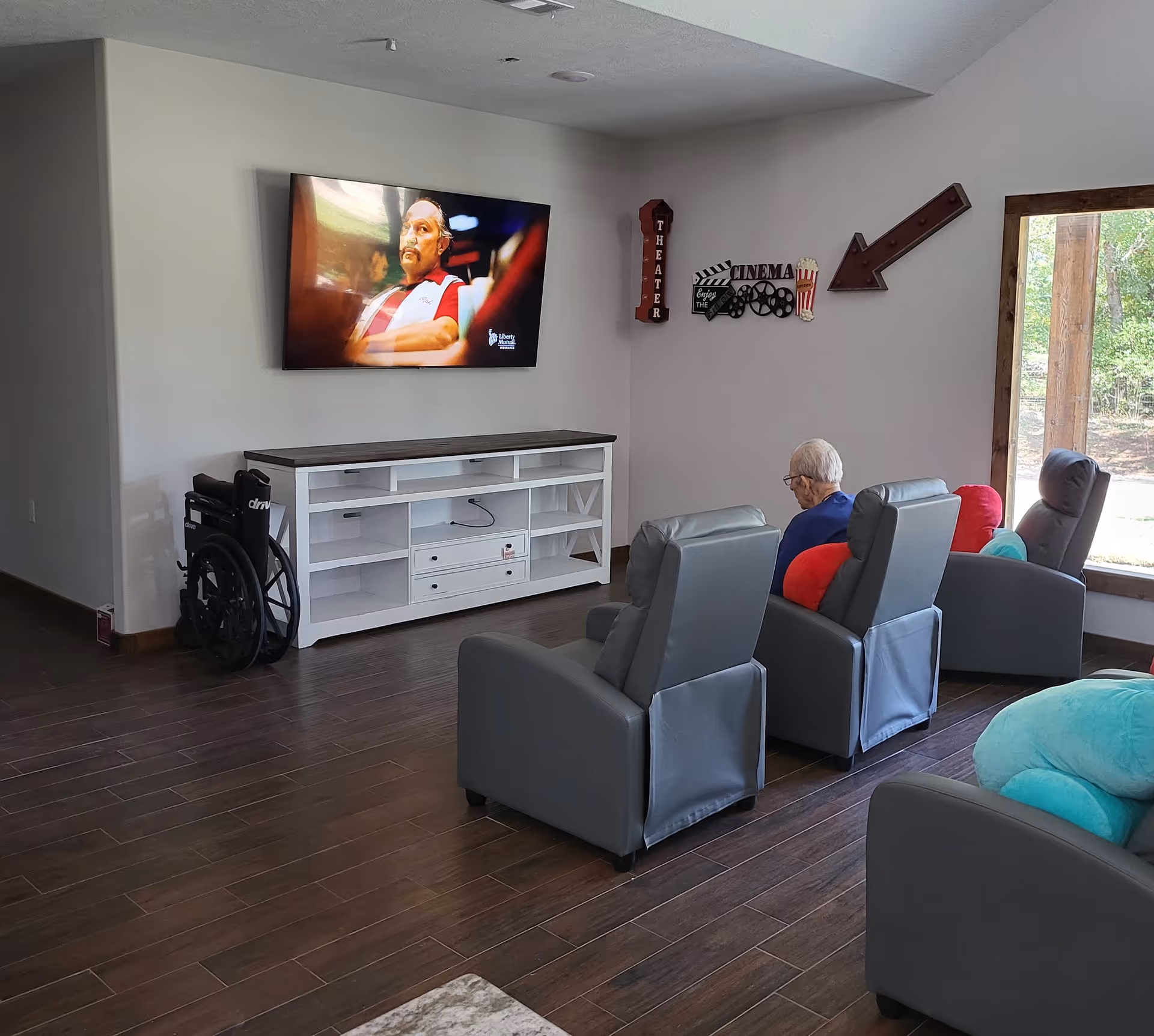 A senior living facility common area with three gray recliner chairs facing a wall-mounted flat screen TV. An elderly person is seated in one of the chairs. There is a white entertainment console below the TV and a wheelchair parked beside it. The wall has decorative signs related to a theater and cinema, and a large window shows greenery outside.