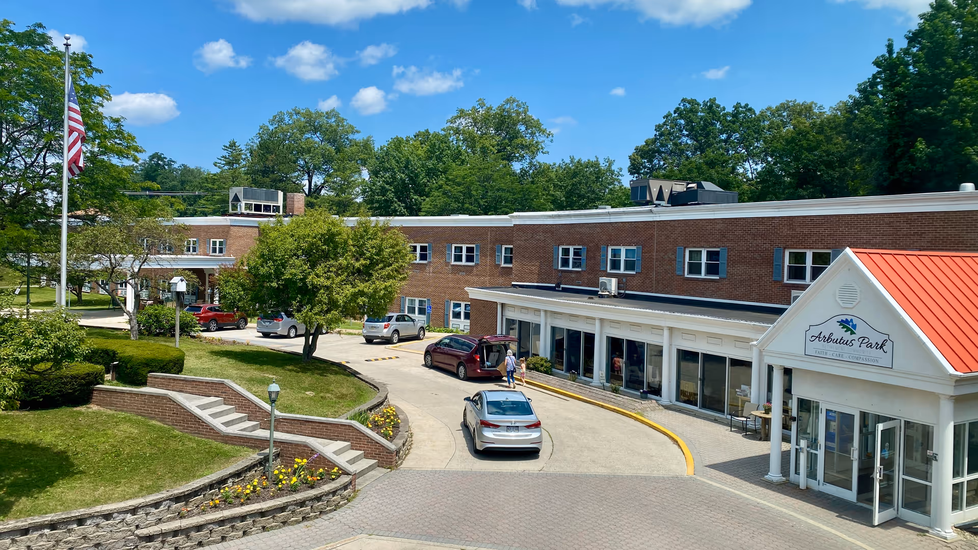 Exterior view of Arbutus Park Retirement Community building with a brick facade and a red roof entrance. Several cars are parked near the entrance, and a person is walking towards the building. The area is surrounded by green trees, bushes, and a well-maintained lawn under a blue sky with scattered clouds.
