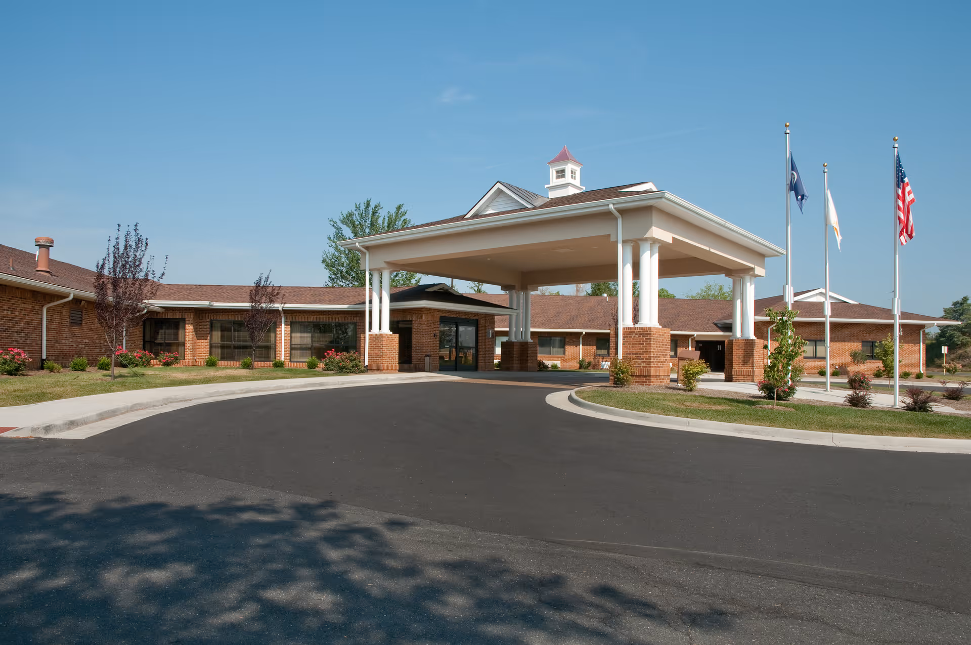 Front entrance of a single-story brick senior living facility with a covered porte-cochere and flagpoles.