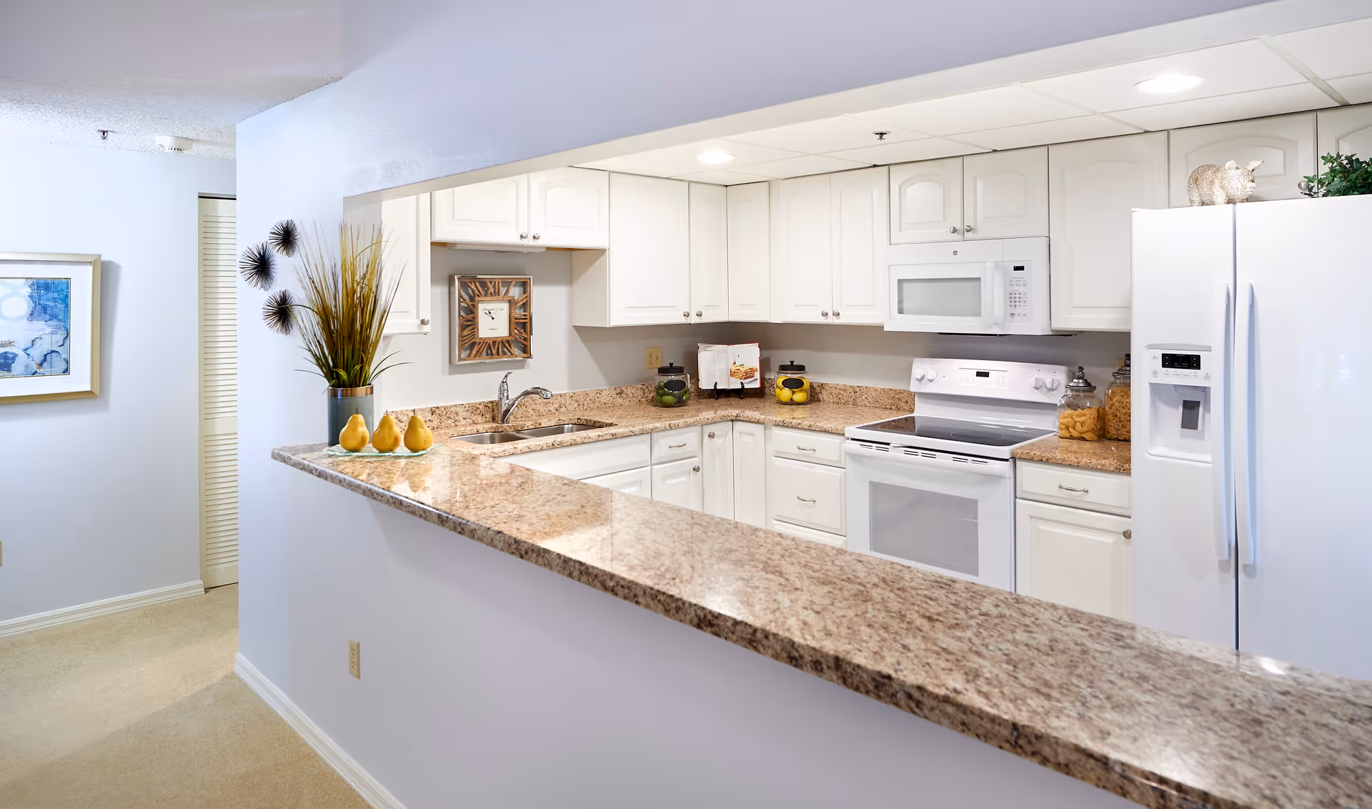 Bright and clean kitchen with white cabinets, a white refrigerator, stove, and microwave. The kitchen features a granite countertop with a sink, decorative items including a vase with tall grass and three pears on the counter, and jars with lemons and pasta on the countertop. A framed picture and a clock are mounted on the wall.