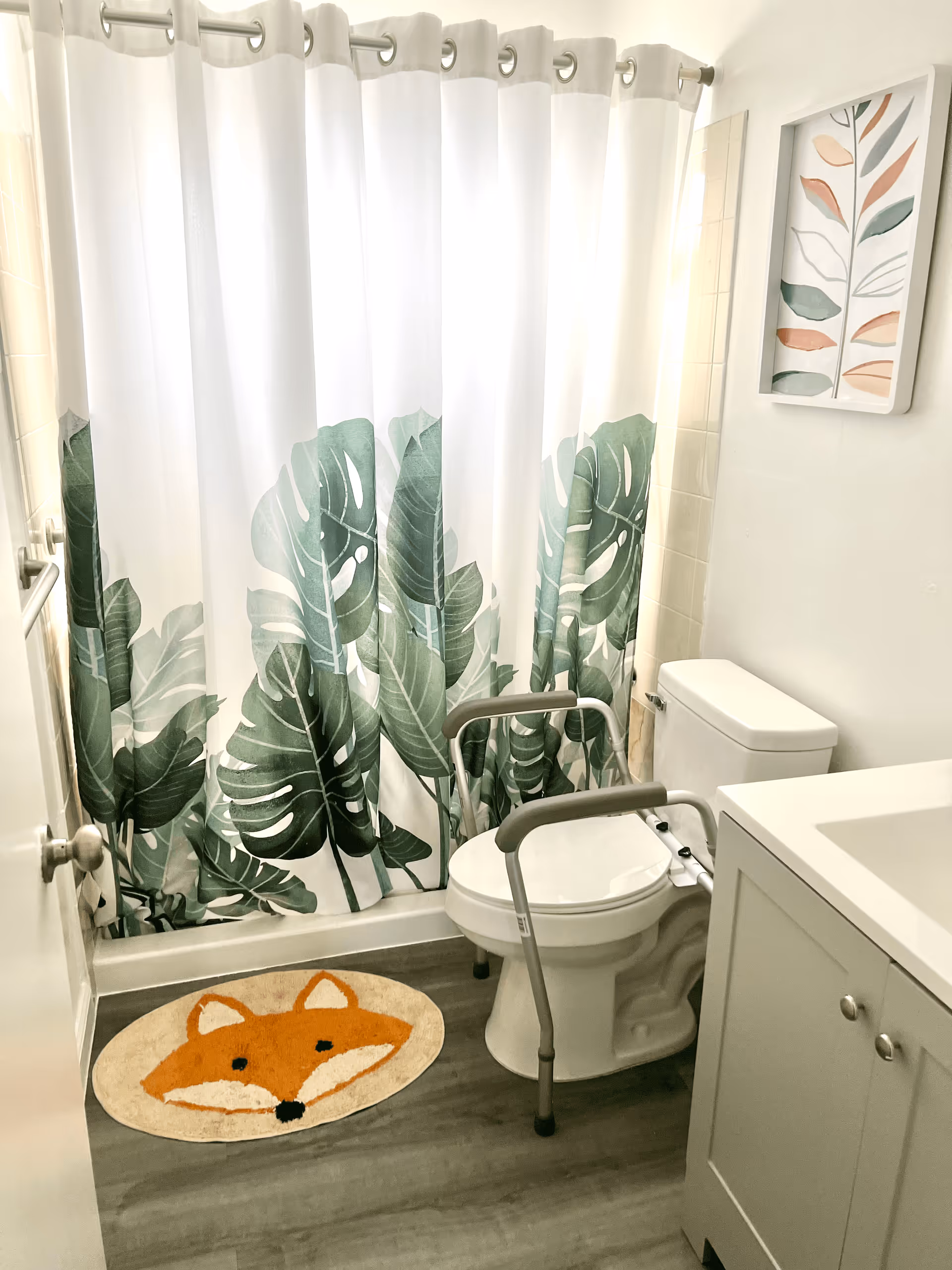 A small bathroom with a white toilet equipped with a safety frame, a white vanity with a sink, a shower with a curtain featuring large green leaves, a round rug with a fox face design on the floor, and a framed artwork of abstract leaves on the wall.