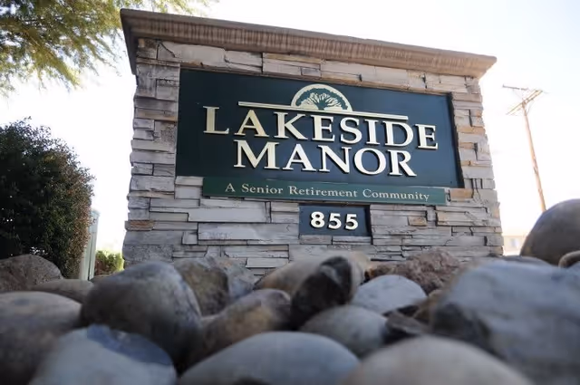 Stone sign for Lakeside Manor, a senior retirement community, with the address number 855 displayed below the name. The sign is surrounded by rocks and some greenery is visible in the background.