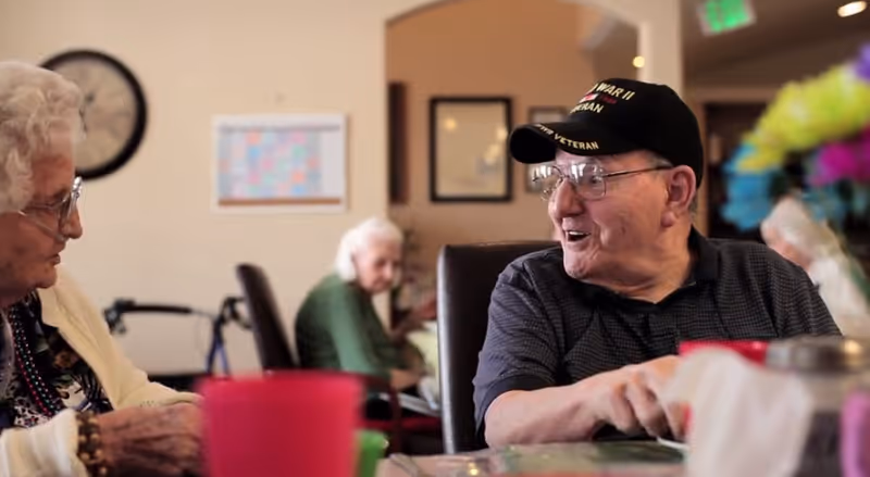 Two older adults converse at a communal dining table in a senior living facility, one man wearing a veteran cap.