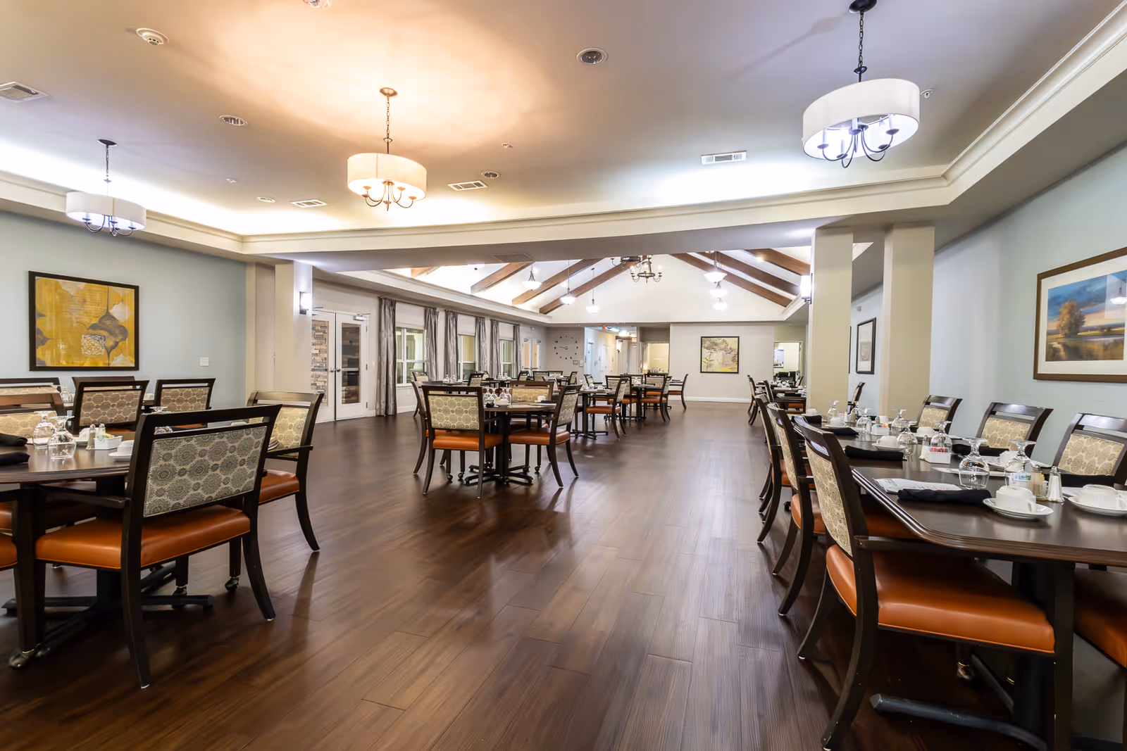 A spacious dining room in an assisted living facility with multiple wooden tables and chairs arranged neatly. The tables are set with cups, glasses, and napkins. The room has wooden flooring, light-colored walls adorned with framed artwork, and ceiling lights providing warm illumination. Large windows with curtains line one side of the room.