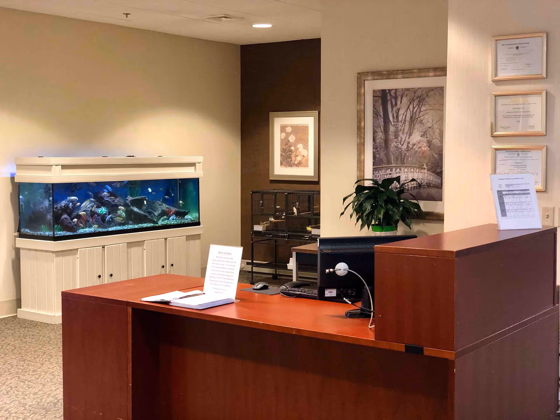 Reception desk area with a computer monitor, a plant, framed certificates on the wall, and a large aquarium filled with fish in the background. The space has neutral-colored walls and carpeted flooring.