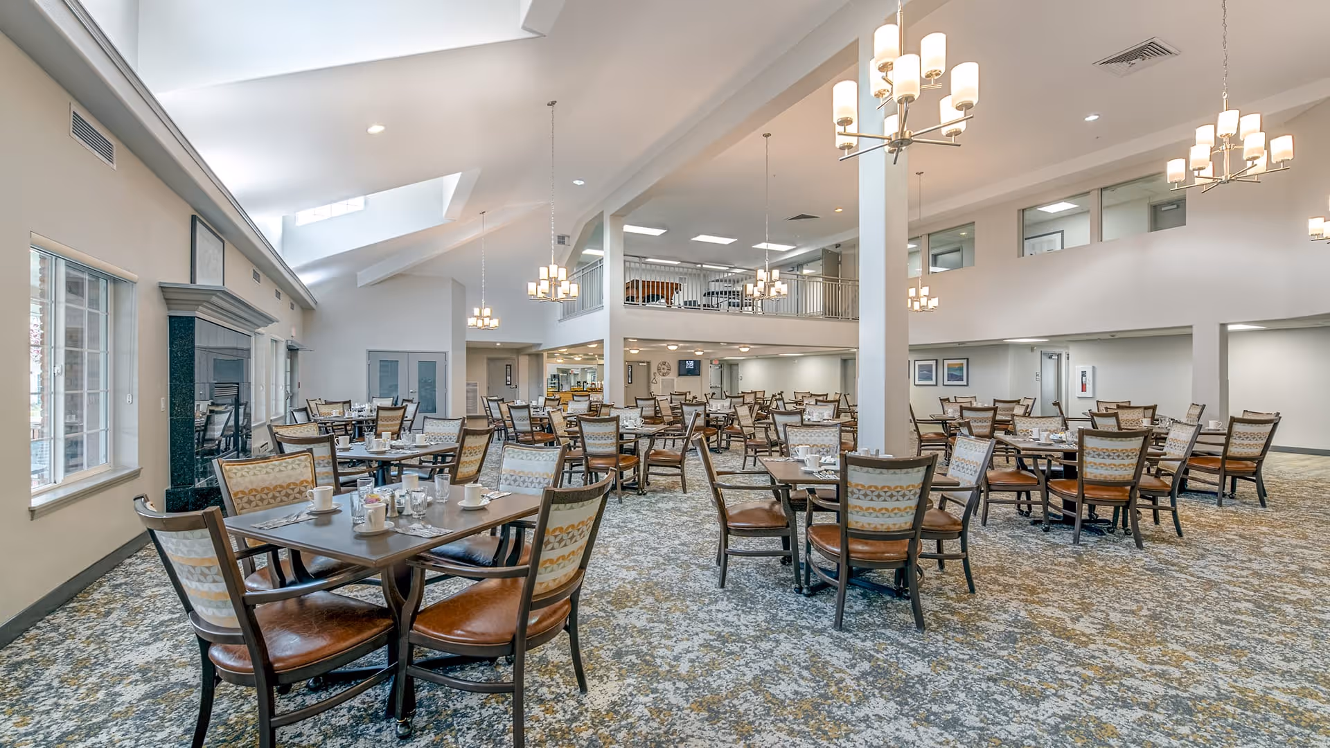 Spacious dining room in a senior living facility with multiple tables and chairs arranged neatly. The room features high ceilings with modern chandeliers, large windows allowing natural light, and a patterned carpet floor. There is a fireplace on one side and a mezzanine level overlooking the dining area.