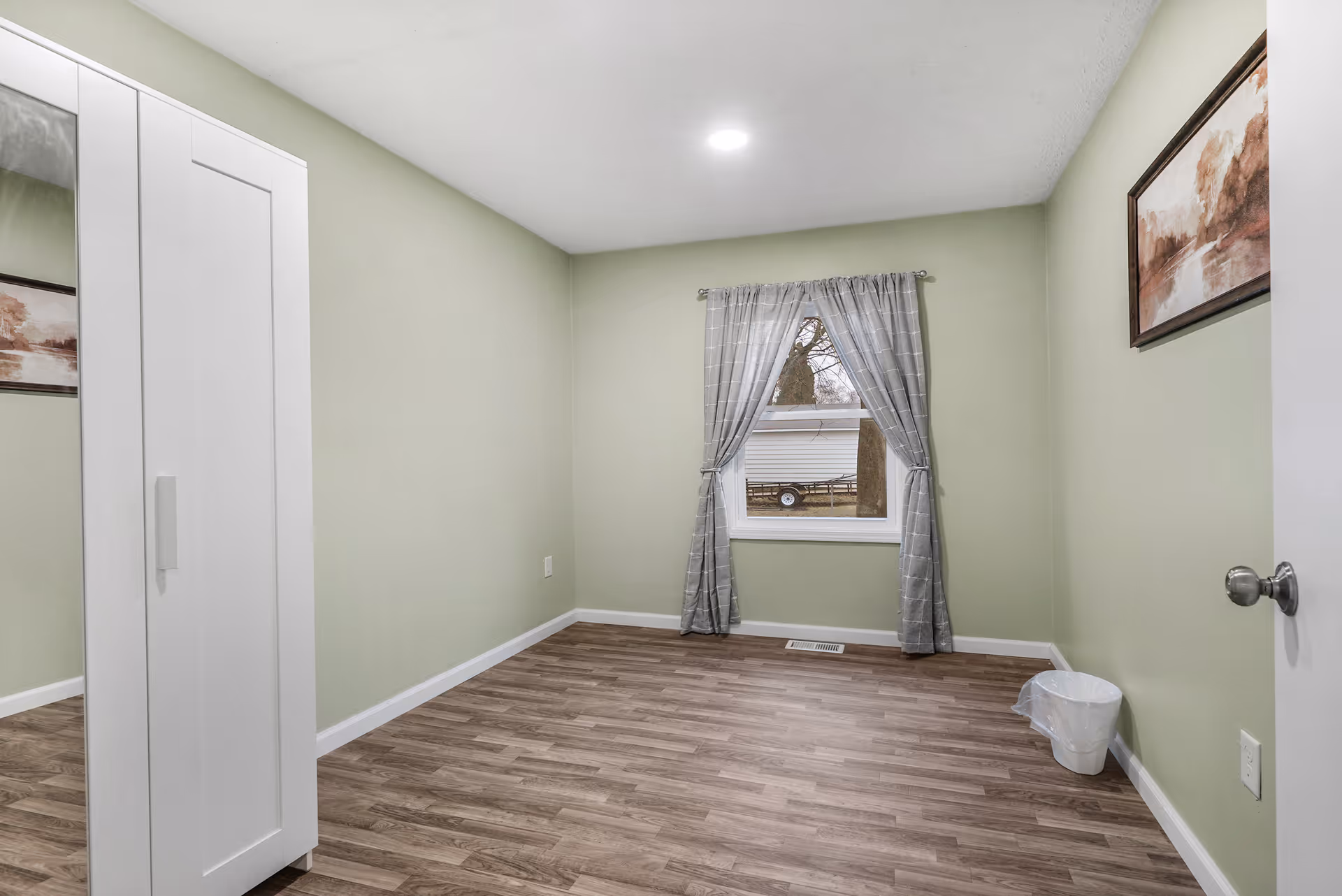 Empty small bedroom with light green walls, wood-look flooring, a window with gray curtains, a white wardrobe and wall art.