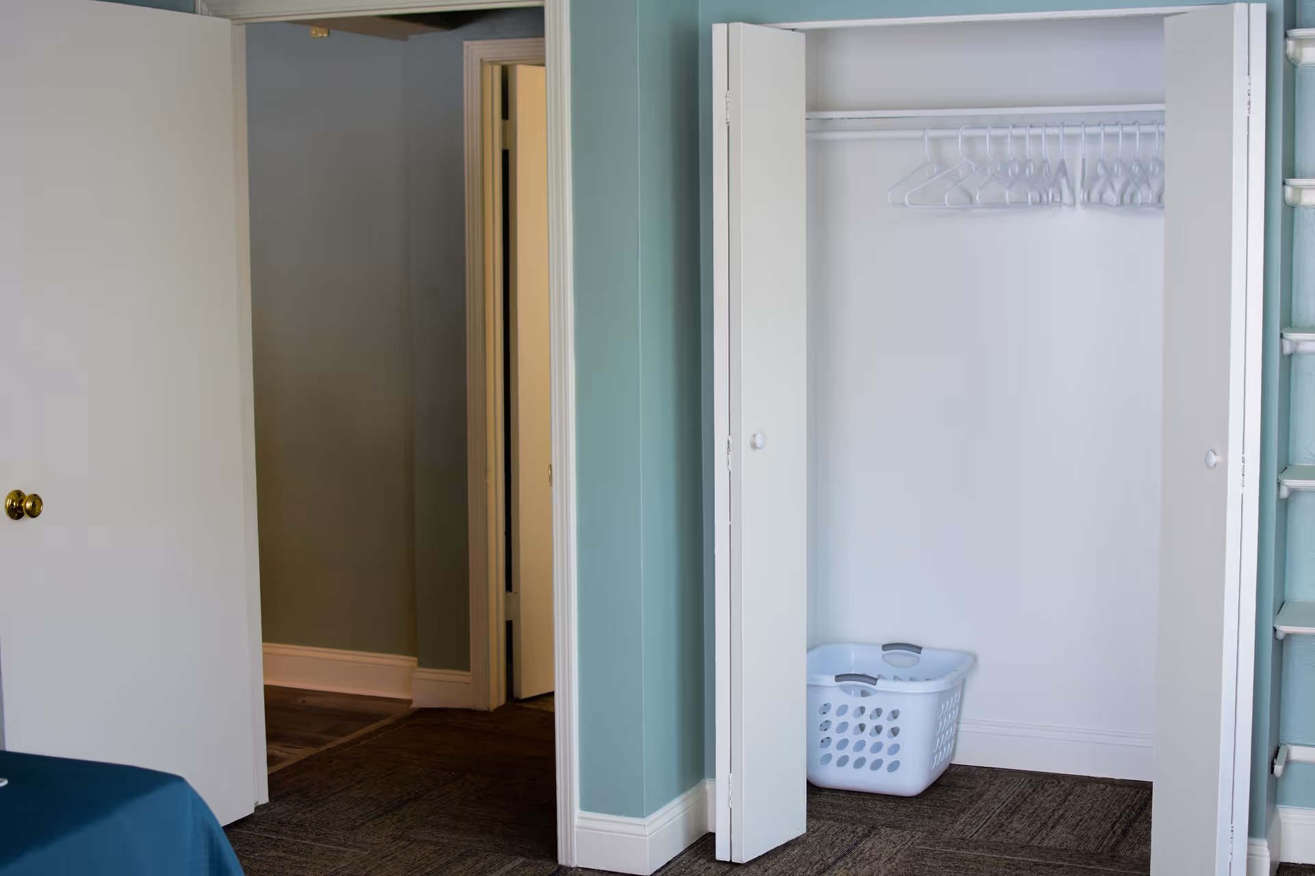 An interior view of a room with light blue walls, an open closet containing several empty white hangers and a white laundry basket on the floor. There is an open door leading to a hallway with wooden flooring and white trim.