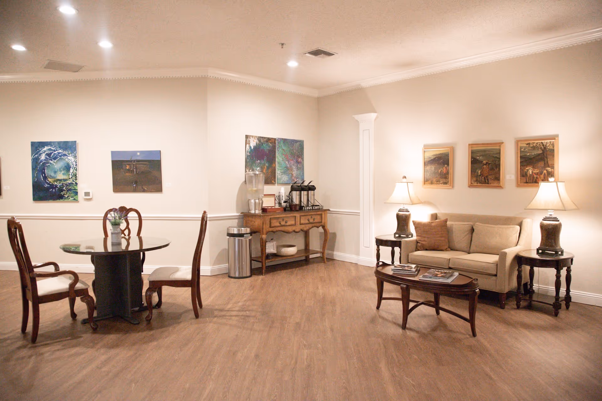 A cozy common area with a beige sofa flanked by two side tables with lamps, a wooden coffee table with magazines, a round dining table with four chairs, and a wooden console table with a water dispenser and coffee station. The walls are adorned with various paintings and the floor is wooden.