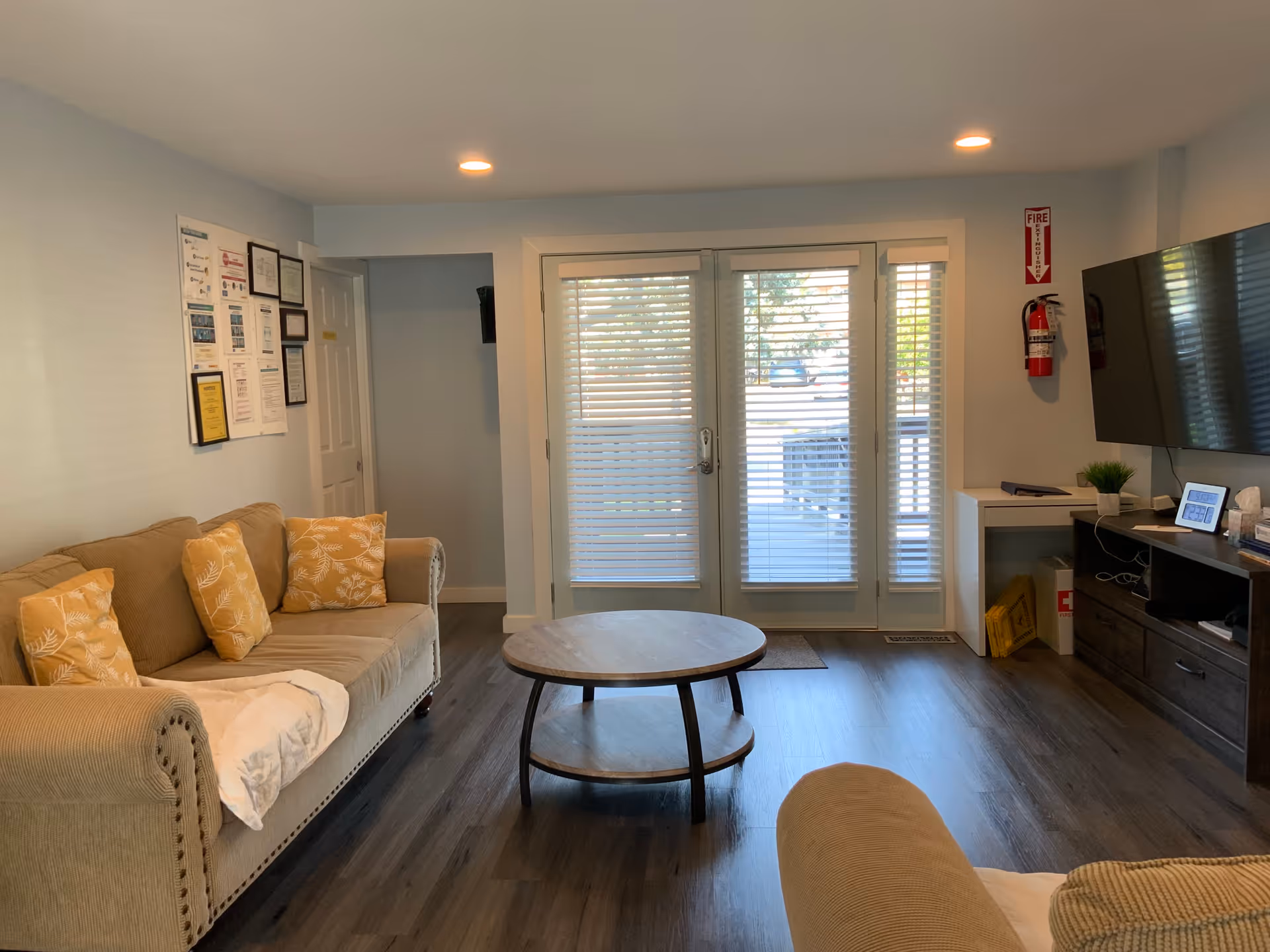 A cozy living room with a beige sofa adorned with yellow patterned pillows, a round wooden coffee table, and a large flat-screen TV mounted on the wall. There are framed certificates and notices on the wall to the left, a fire extinguisher mounted on the wall near the TV, and glass doors with blinds leading outside.