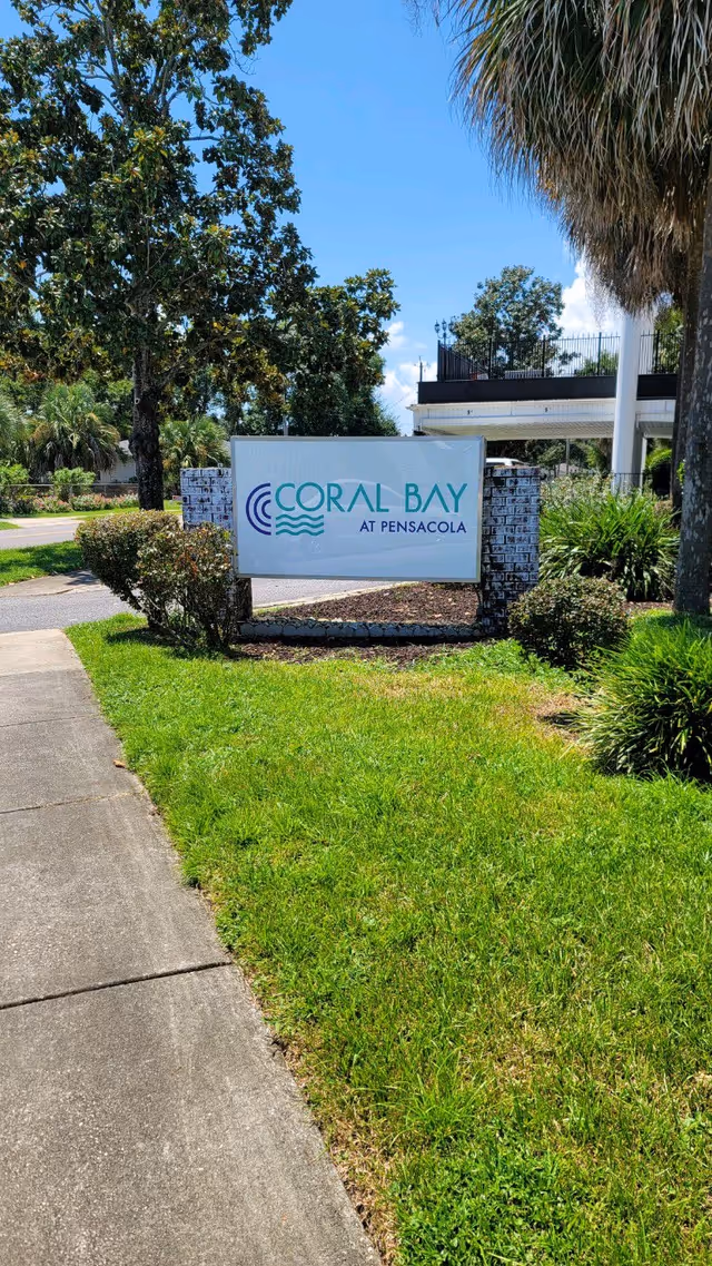 Outdoor entrance sign that reads "Coral Bay at Pensacola" on a landscaped lawn with trees and part of the building in the background.