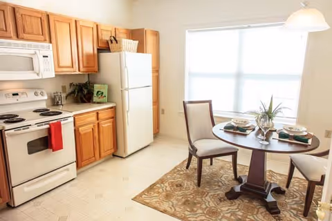 A bright kitchen and dining area featuring wooden cabinets, a white refrigerator, stove, and microwave. A round wooden dining table with two chairs is set with plates, glasses, and napkins, placed on a patterned rug near a large window with blinds.