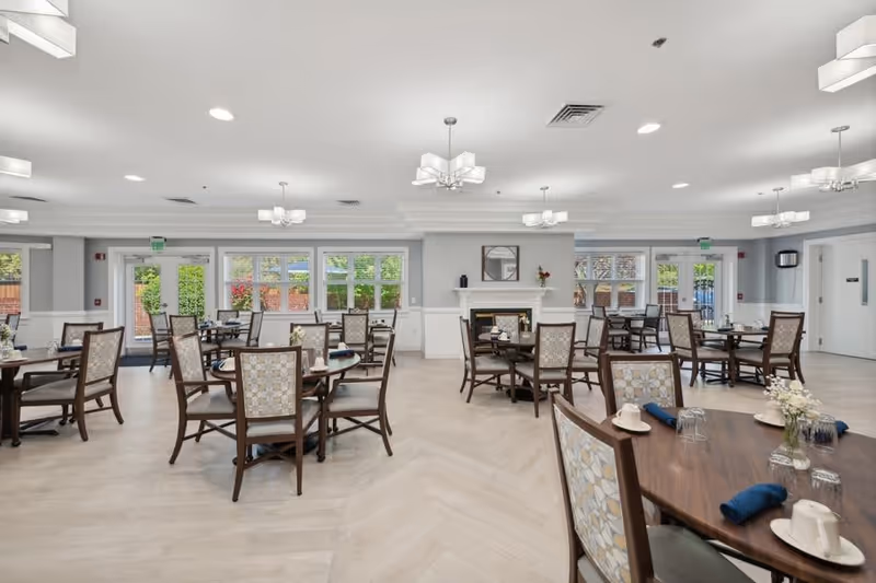 A bright and spacious dining room in a senior living community with multiple round wooden tables and cushioned chairs arranged neatly. Each table is set with cups, glasses, and napkins. Large windows and glass doors allow natural light to fill the room, and a fireplace with a mirror above it is visible on one wall. Modern ceiling lights hang from the white ceiling.