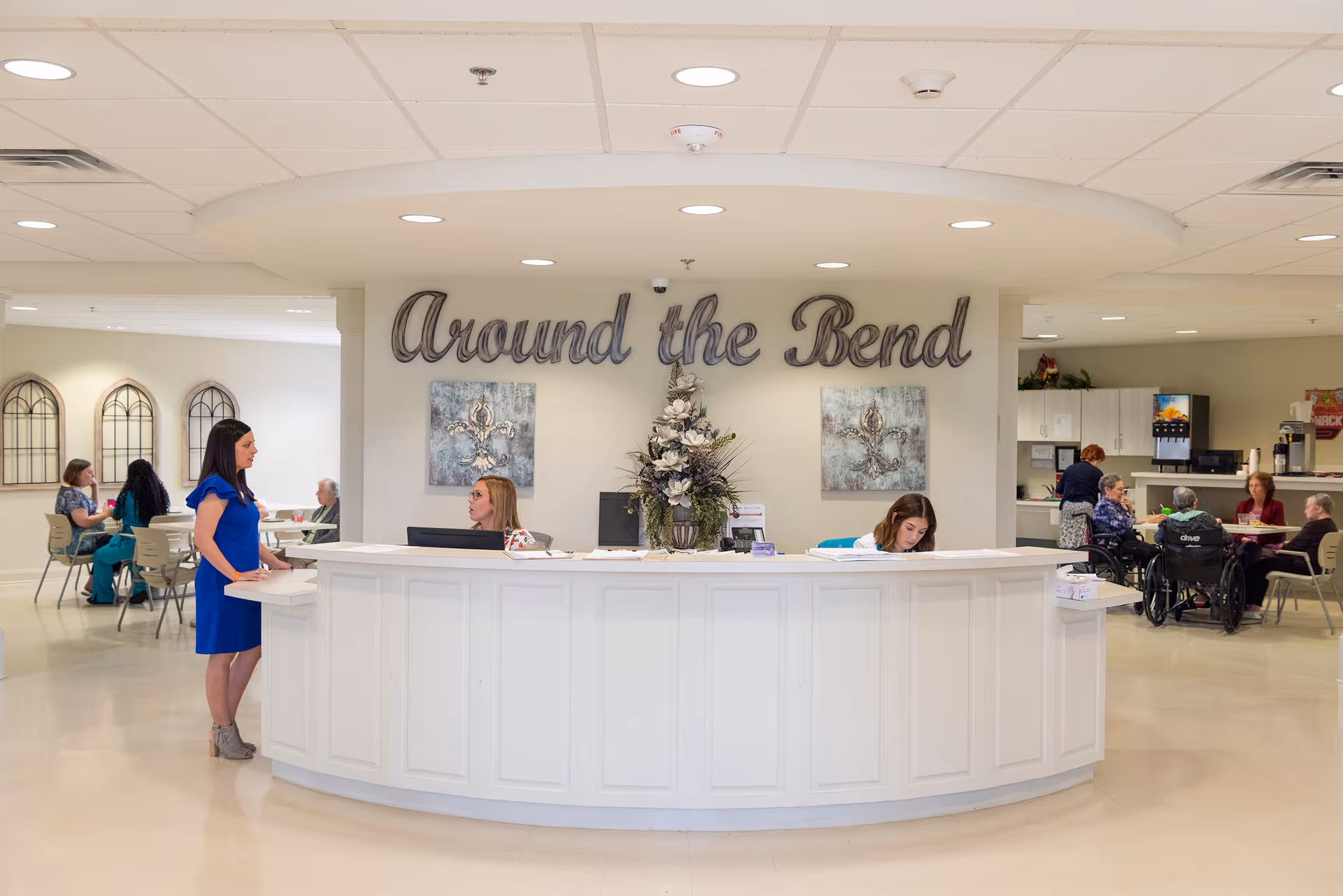 Bright senior living lobby with a curved white reception desk, staff and residents nearby, and a wall sign reading "Around the Bend."