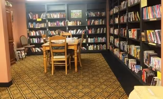Carpeted common room with wall-to-wall bookshelves and a wooden table with chairs in the center.