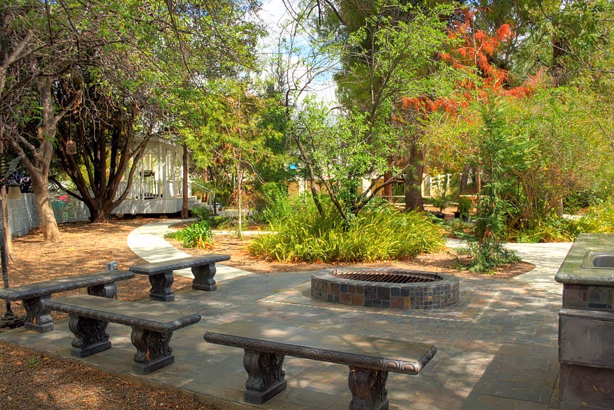 Outdoor garden area with stone benches arranged around a circular fire pit, surrounded by trees and greenery. A paved walkway curves through the garden, and a small white gazebo is visible in the background.