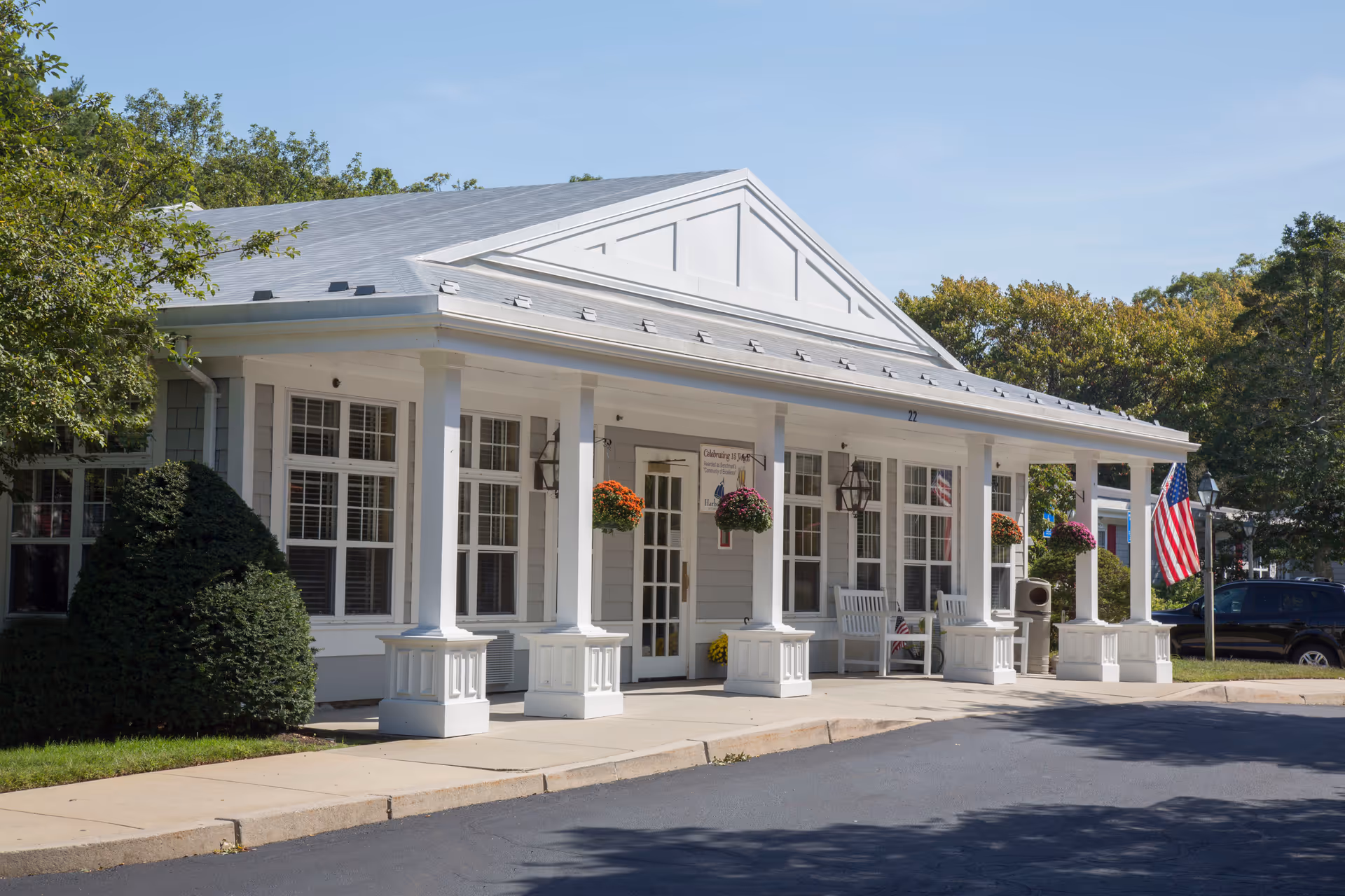 Exterior view of a single-story building with white columns and a gray roof, featuring hanging flower baskets and American flags near the entrance. The building is surrounded by trees and bushes under a clear blue sky.