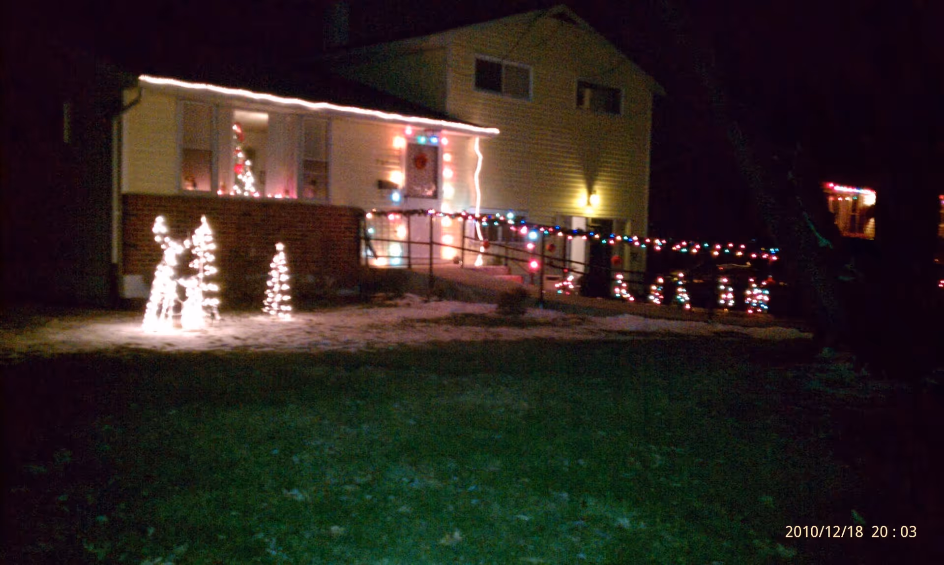 Night view of a house decorated with Christmas lights including illuminated reindeer figures, a small Christmas tree, and multicolored string lights along the railing and roofline. Snow is visible on the ground.