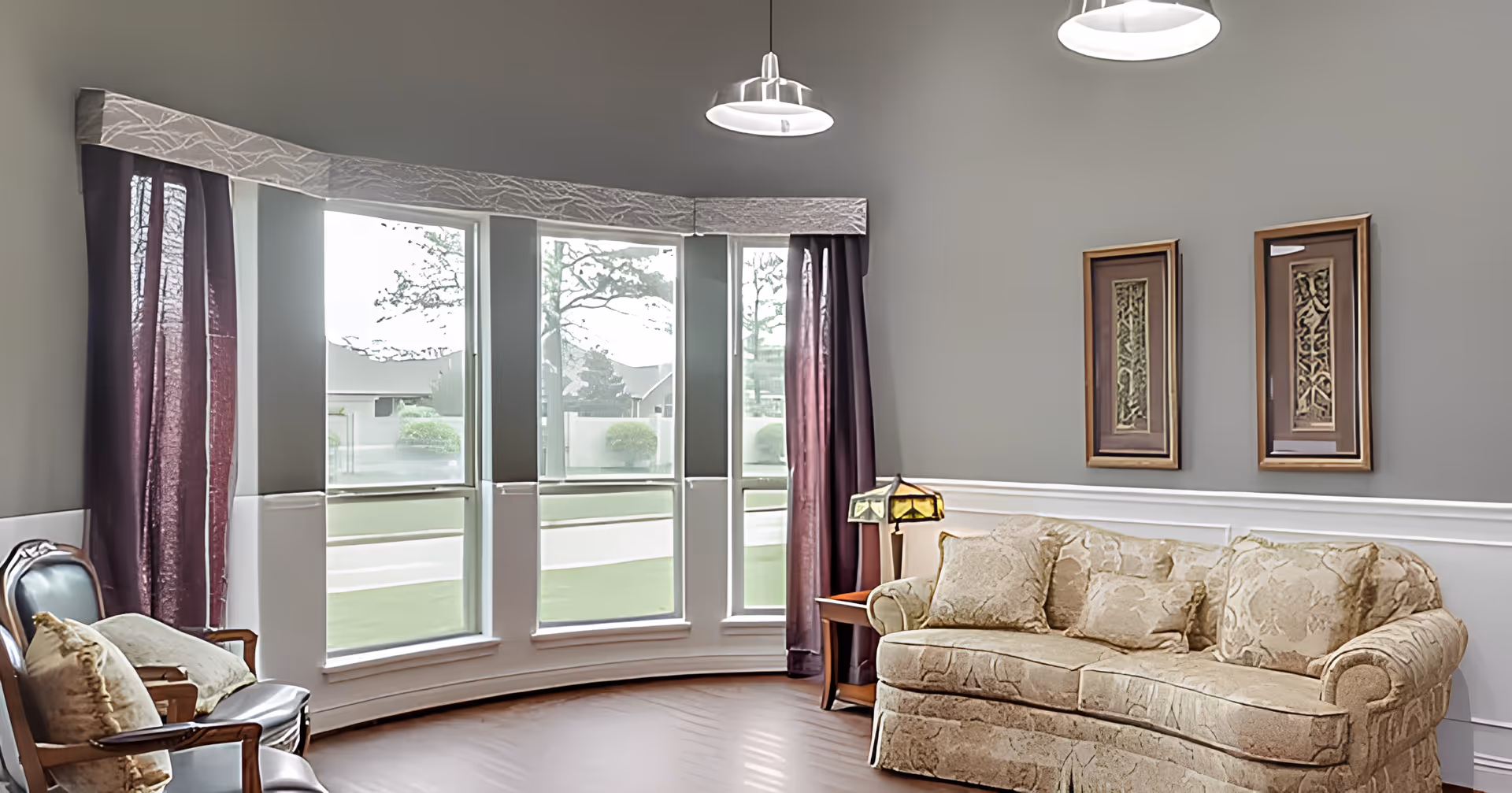 A cozy living room with a beige patterned sofa and two wooden chairs with cushions. The room features a large curved window with sheer curtains and dark purple drapes. Two framed artworks hang on the gray wall above the sofa. A small side table with a decorative lamp is placed next to the sofa. The floor is wooden, and two ceiling lights are visible.