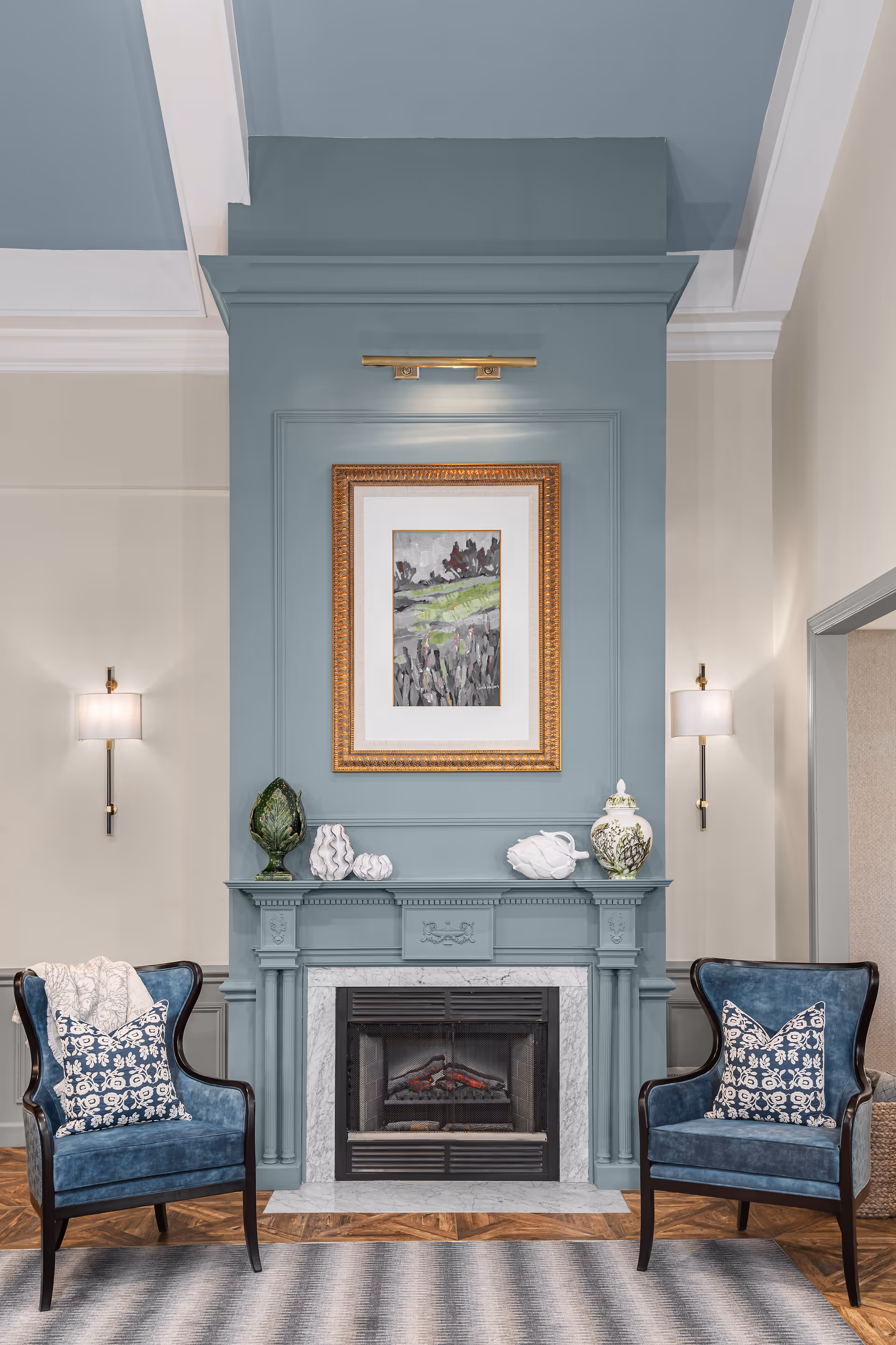 A cozy living room area featuring a blue fireplace mantel with a marble surround. Above the fireplace is a framed painting of a landscape with green and gray tones, illuminated by a brass picture light. On the mantel are decorative items including a green artichoke sculpture, white coral pieces, and a white vase with green floral designs. Flanking the fireplace are two blue upholstered armchairs with dark wood frames, each adorned with a patterned cushion. The walls are painted in soft beige and blue tones, with two wall sconces providing additional lighting. The floor has a wood pattern and a gray area rug.