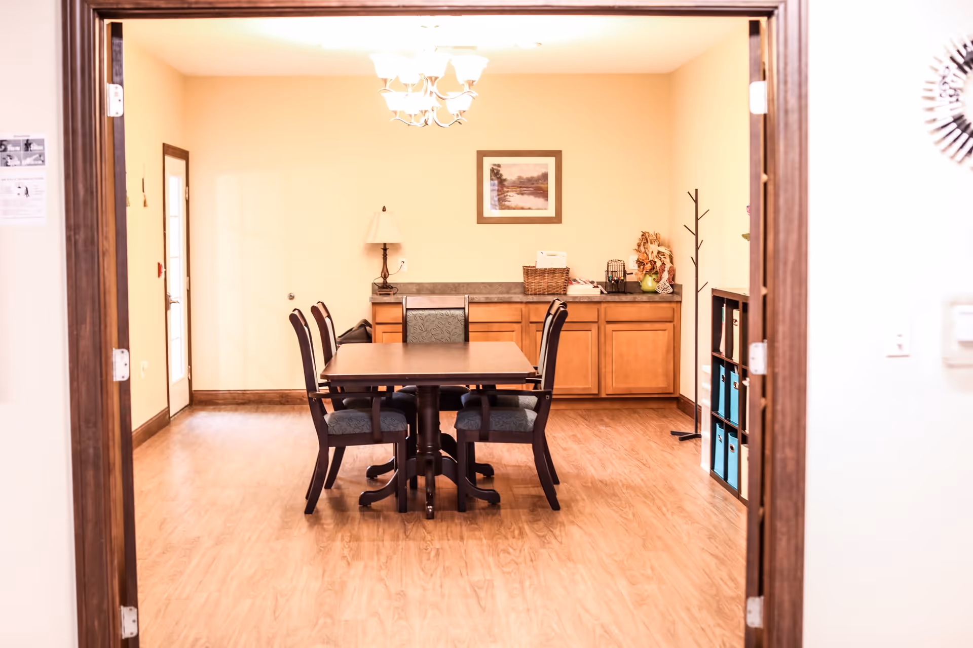 Communal dining room with a wooden table and chairs, cabinets, lamp and wall art seen through a doorway.