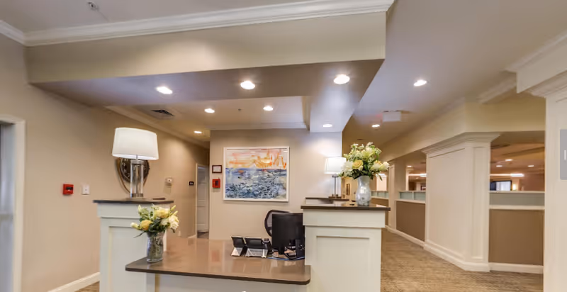 Reception desk in a well-lit lobby with lamps, flowers, artwork, and a computer.