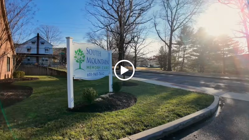 Sign for South Mountain Memory Care on a grassy lawn beside a road, with trees and a building in the background.