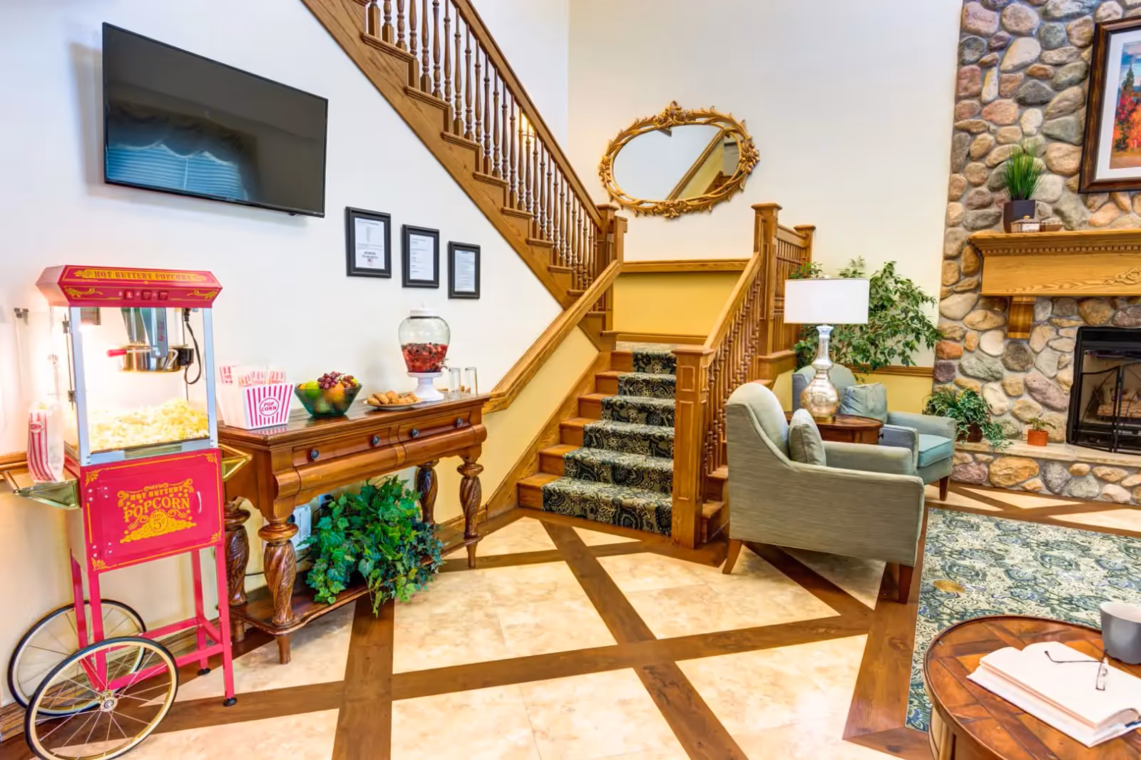 A cozy interior living area featuring a wooden staircase with a patterned carpet runner, a stone fireplace with a wooden mantle, two upholstered armchairs with a small table and lamp between them, a wooden console table with snacks and a beverage dispenser, and a red vintage-style popcorn machine. A flat-screen TV is mounted on the wall above the console table, and a decorative oval mirror hangs above the staircase landing.