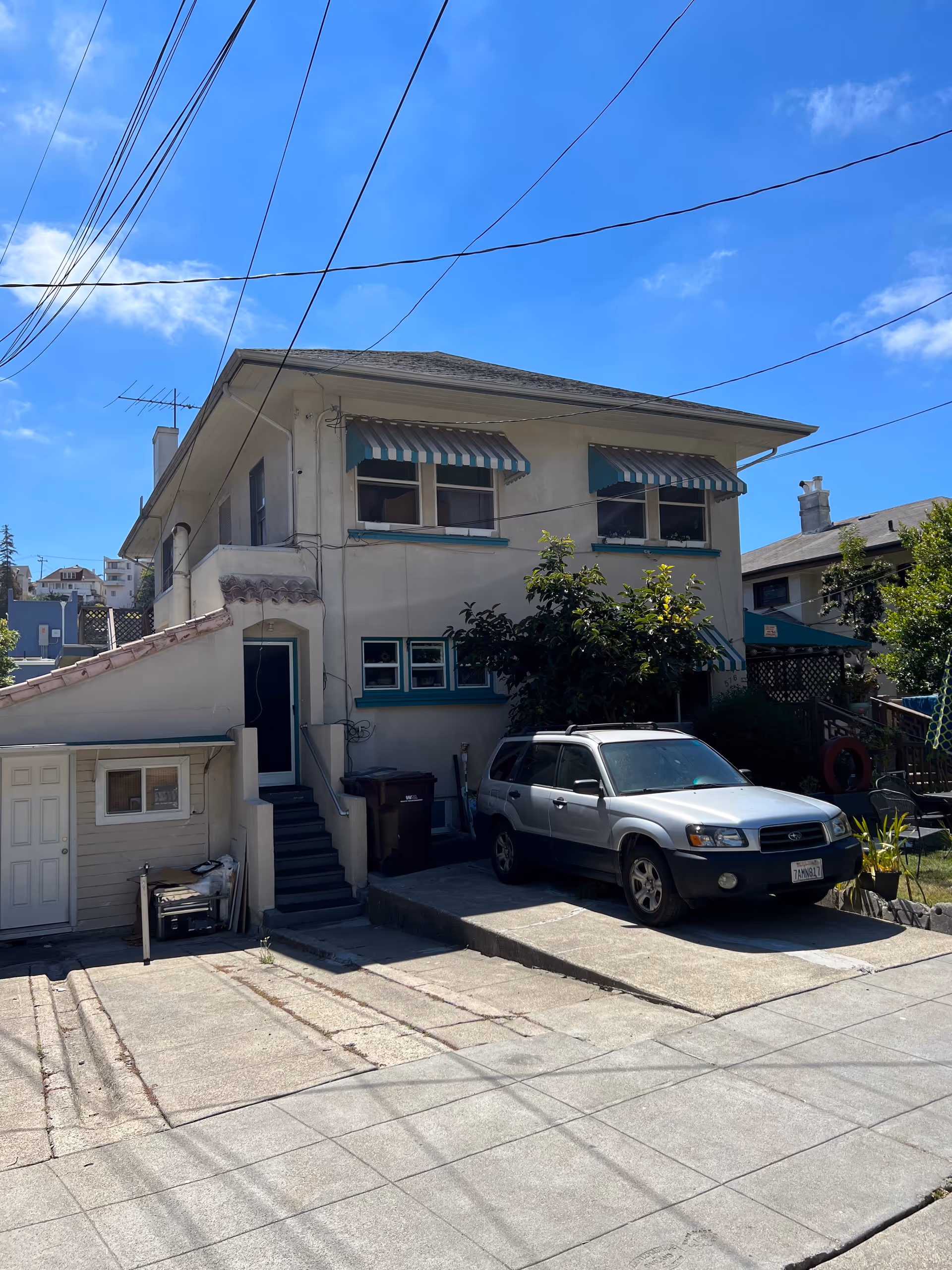Exterior view of a two-story residential building with striped awnings over the windows, a parked silver SUV in the driveway, and a clear blue sky above.