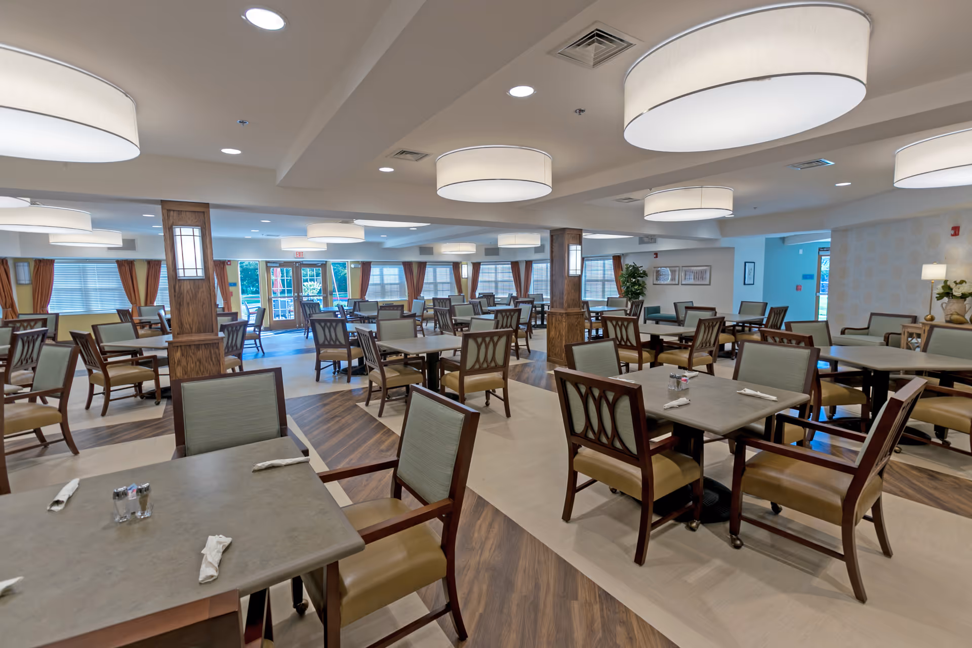 Spacious dining room with multiple tables and chairs arranged neatly. The room features large circular ceiling lights, wooden pillars, and windows with orange curtains allowing natural light to enter. The floor has a combination of wood and tile patterns.