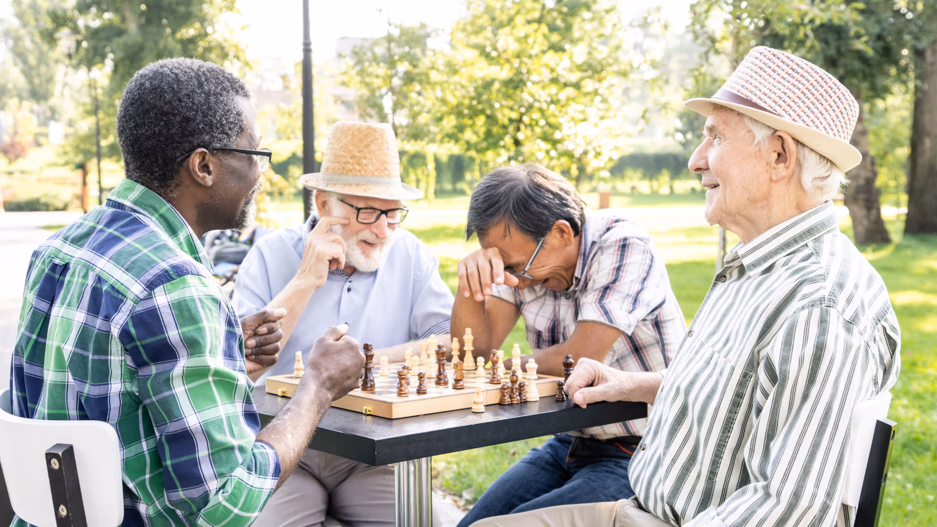 Four elderly men sitting outdoors around a table playing chess on a sunny day in a park-like setting with green trees and grass in the background. They appear engaged and happy, with one man laughing and another smiling.