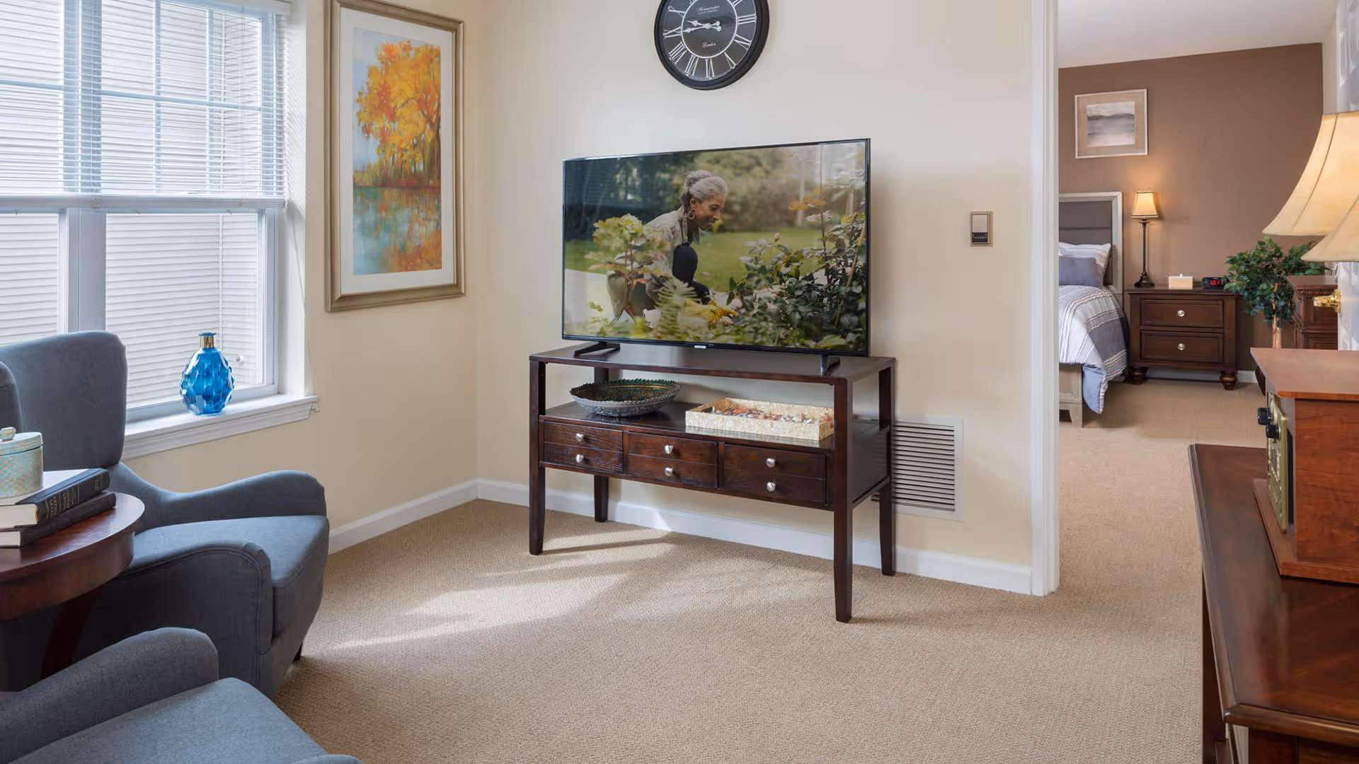 A cozy living room area with two gray armchairs, a wooden side table with books and a decorative jar, a flat-screen TV on a wooden console table, and a large window with blinds. A colorful painting hangs on the wall next to the TV. In the background, a bedroom with a bed, nightstand, lamp, and framed artwork is visible through an open doorway.