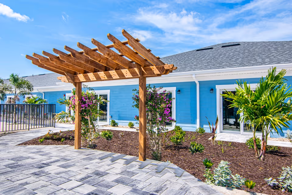 Outdoor area of Hampton Manor of Cape Coral featuring a wooden pergola over a paved walkway, surrounded by landscaped garden beds with various plants and flowers, and a bright blue building with white trim in the background under a partly cloudy sky.