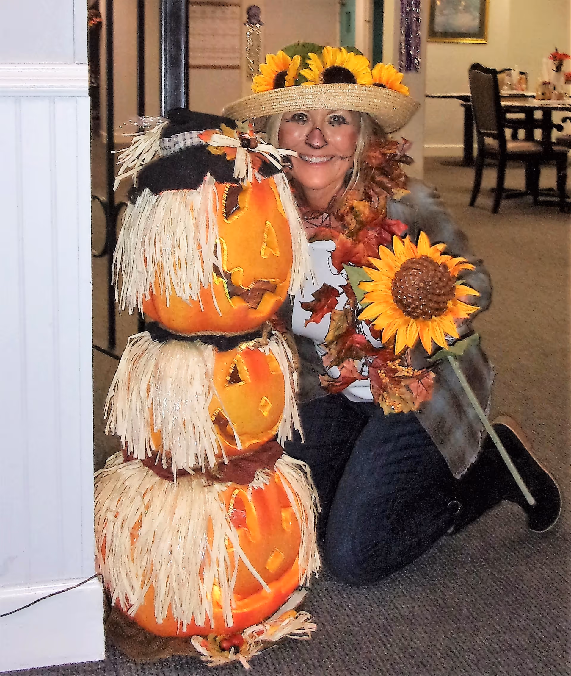 A person dressed in a festive autumn costume with a straw hat decorated with sunflowers, face painted with a nose and whiskers, kneeling next to a stacked pumpkin decoration with carved faces and straw accents, holding a large artificial sunflower. The setting appears to be an indoor common area with tables and chairs in the background.
